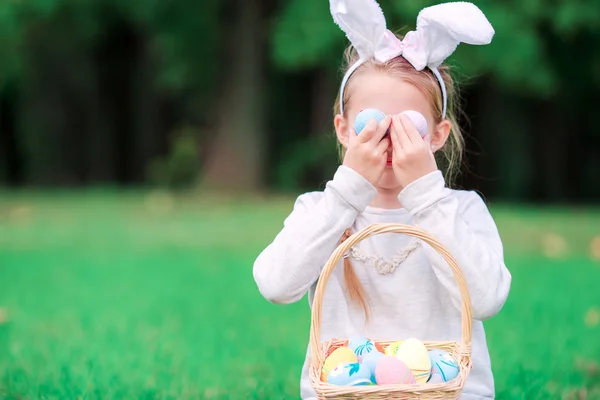 Lustiges kleines Mädchen mit Hasenohren, das an einem Frühlingstag draußen mit Ostereiern spielt — Stockfoto