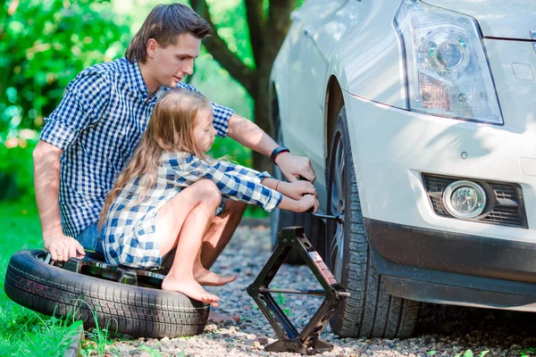 Adorable little girl sitting on a tire and helping father to change a car wheel outdoors on beautiful summer day