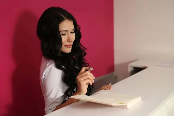 A beautiful woman female in a white uniform stands reception and writes with a pen in a notebook. Modern design of the premises of the beauty salon of the office of medical clinic. Administrator