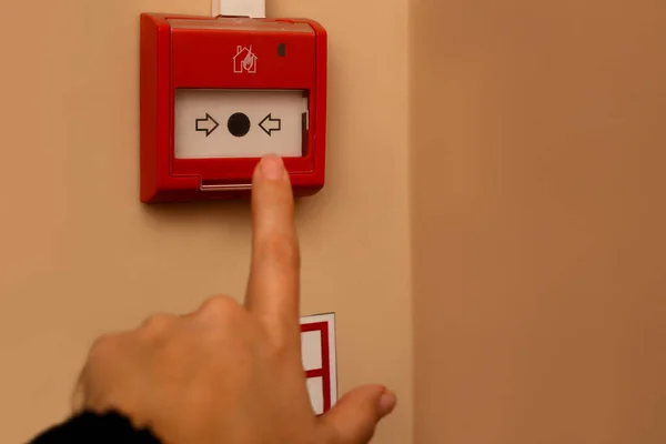 A kéz ujja kinyúlik, hogy megnyomja a piros riasztó gombot a tűzjelző a falon. Figyelem, evakuálás! Kifogyott a kültéri — Stock Fotó