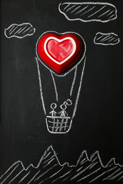 恋人同士が熱気球に乗って飛んでいる。黒い黒板に白いチョークで大きな赤いハートの絵。バレンタインデーのためのロマンチックな写真。お祝いのポストカード. — ストック写真