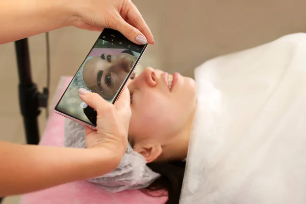 Permanent makeup skönhetssalong mästare tar ett foto på mobilen av kunderna ansikte efter att ha avslutat sitt arbete — Stockfoto