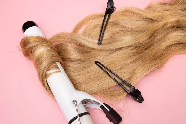 Curling blonde hair on a large diameter curling iron on a pink background. Curl care, hair styling, black clips.