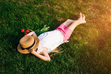Hasır şapkalı bir kız güneşli bir yaz gününde parkta çimlerin üzerinde dinleniyor. Sırtüstü yatar, şapka yüzünü kapatır..