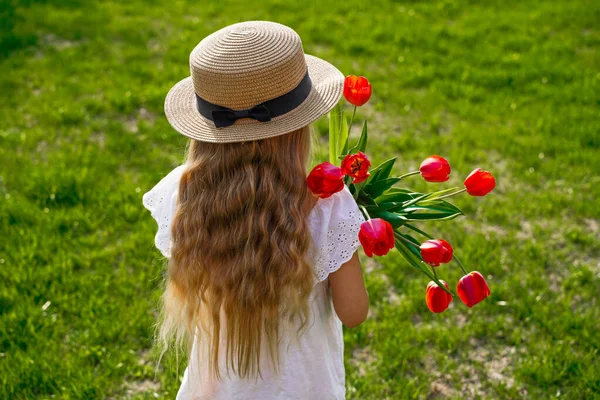 帽子に長いブロンドの黄金の波状の髪を持つ子供の女の子の背中からのバックビュー、緑の芝生の背景に対して、赤いチューリップの花束を保持. — ストック写真