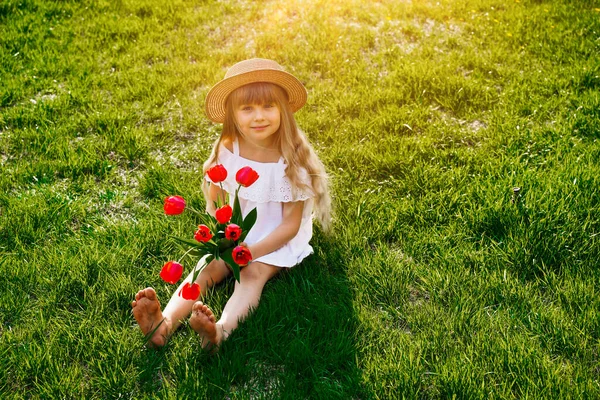 Smiling happy child girl with long blond golden wavy hair in a hat, against a background of green grass lawn, holding a bouquet of red tulips. — Stock Photo, Image