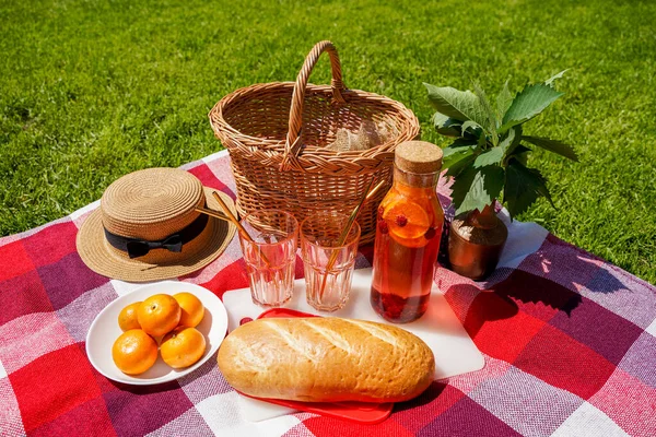 Piknik na trávě, červenou kostkovanou přikrývku, proutěný koš, přírodní nápoj z pomerančů a bobulí, bochník. Lahodné občerstvení venku v létě — Stock fotografie