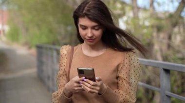 Sosyal medyada sohbet eden güzel bir kadının fotoğrafını çek. Uzun saçlı, neşeli esmer kadın. Şehir parkında yürürken mesaj yazıyor ve gülümsüyor..
