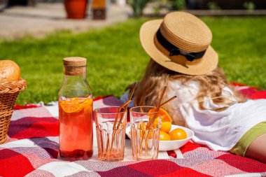 Çimlerin üzerinde piknik yapan bir kız, kırmızı bir ekose battaniye, hasır bir sepet, portakal ve böğürtlenlerden yapılmış doğal bir içecek, bir somun ekmek. Yazın dışarıda nefis atıştırmalıklar.