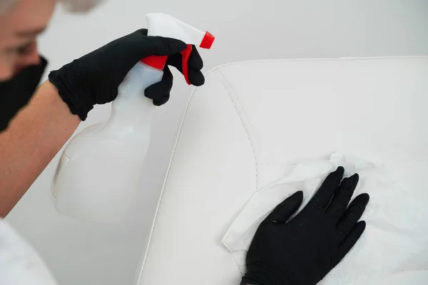 Pulizia del posto di lavoro nella SPA, salone di bellezza. Una giovane donna con una maschera protettiva e guanti tiene in mano uno spray antisettico, pulisce la sedia dei clienti con un tovagliolo. — Foto Stock