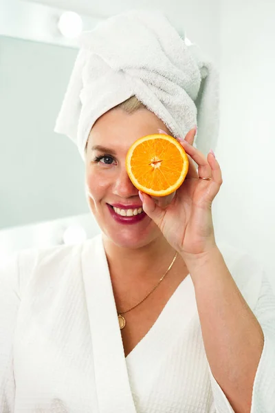 Portrait de beauté d'une jolie femme joyeuse en robe blanche avec une serviette enroulée autour de son visage tenant des tranches d'orange sur le visage et regardant la caméra dans la salle de bain blanche — Photo