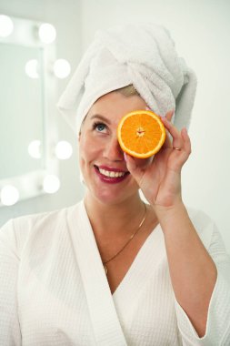 Banyoda beyaz bornozlu bir kadın elinde portakalın iki yarısını tutuyor, cilt bakımı, yüz için vitamin, enerji ve sağlık.
