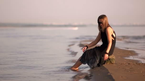 Fille dans une robe noire assis sur un rocher au bord de la rivière pieds dans l'eau — Video