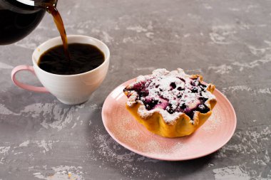 Taze sade kahve fincana doldurulur. Süzme peynir ve böğürtlenli kek sepeti, üzerine pudra şekeri serpilmiş. Beton bir masanın arka planında kahvaltı.