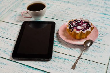 Dijital bir tablet, bir fincan kahve ve beyaz ve mavi renkli bir masanın üzerinde çilek ve süzme peynirli bir kek sepeti. Sabah kahvaltısı.