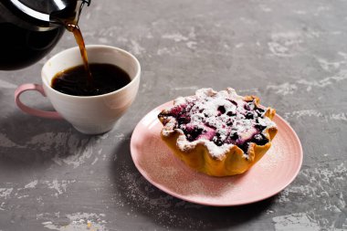 Taze sade kahve fincana doldurulur. Süzme peynir ve böğürtlenli kek sepeti, üzerine pudra şekeri serpilmiş. Beton bir masanın arka planında kahvaltı.