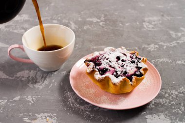Taze sade kahve fincana doldurulur. Süzme peynir ve böğürtlenli kek sepeti, üzerine pudra şekeri serpilmiş. Beton bir masanın arka planında kahvaltı.