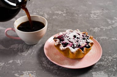 Taze sade kahve fincana doldurulur. Süzme peynir ve böğürtlenli kek sepeti, üzerine pudra şekeri serpilmiş. Beton bir masanın arka planında kahvaltı.