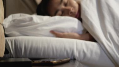 Asyalı kadın uykulu bir şekilde yatakta uyur ve sabah alarmın sesini kapatır..