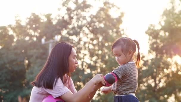 若い母親は 夏の公園でスクーターに乗ることを学んだ後 彼女の娘が保護肘パッドを削除するのに役立ちます 子供のためのアクティブな屋外スポーツ — ストック動画