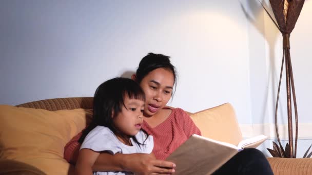 快乐的妈妈抱着可爱的小女儿一起坐在沙发上看书童话故事 睡前故事概念 — 图库视频影像