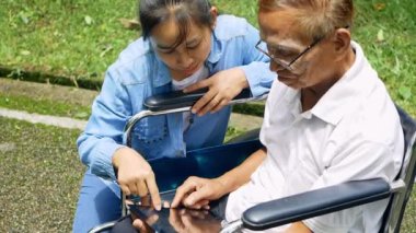 Büyükbaba tekerlekli sandalyede ve torun parkta dijital tabletle eğleniyor. Tatilde aile hayatı.