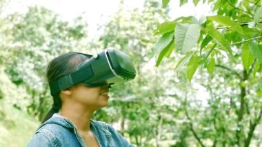 Ormanda sanal gerçeklik gözlüğü takan ve bahar bahçesinde güneşli bir yaz gününde doğanın tadını çıkaran mutlu kadın. Modern teknoloji kavramı.