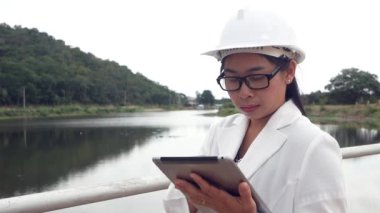 Beyaz miğferli kadın mühendis dijital tabletle çalışıyor ve baraj inşaatına bakıp elektrik üretiyor..