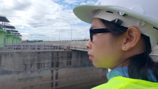 白いヘルメットと黄色の制服の女性エンジニアは青写真を保持し 発電するためにダム建設現場を離れて見て — ストック動画