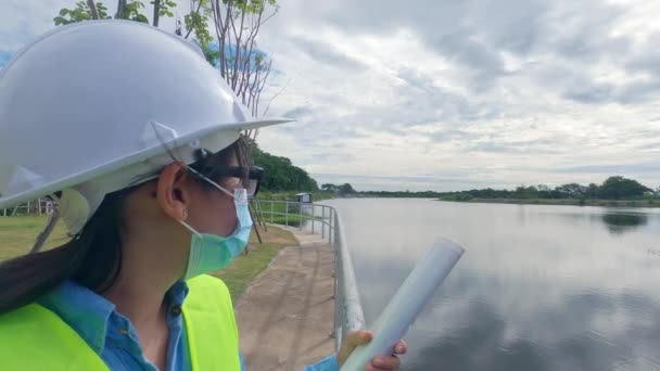 身穿白色头盔和黄色制服 戴着保护面罩的女工程师拿着一个设计图 看着大坝建筑工地以外的地方发电 — 图库视频影像