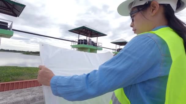 身穿白色头盔和黄色制服的女工程师在图纸上阅读数据 并检查大坝建筑工地的供水系统以发电 — 图库视频影像