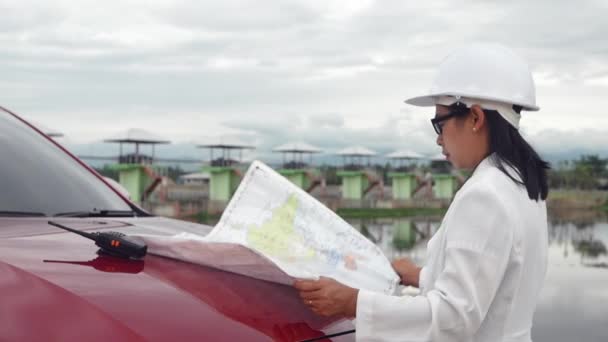 身穿白色头盔的女工程师阅读放置在汽车前盖的蓝图中的数据 并在大坝建筑工地使用无线电通信发电 — 图库视频影像