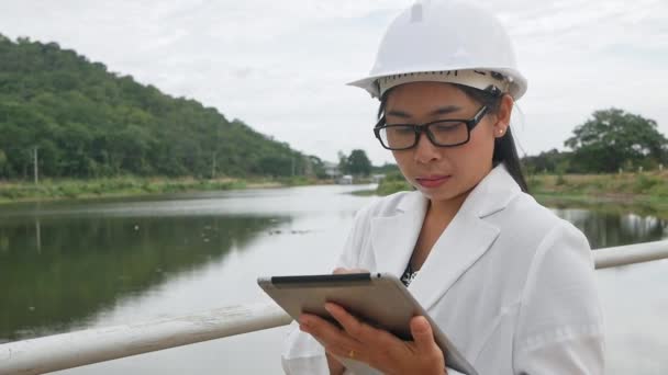 戴白色头盔的女工程师正在使用数字平板电脑 看着大坝建筑工地以外的地方发电 — 图库视频影像