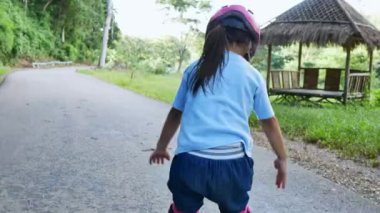 Koruyucu pedler takan ve kasklı küçük bir kız yaz parkında paten kaymayı öğreniyor. Çocuklar için aktif açık hava sporu.