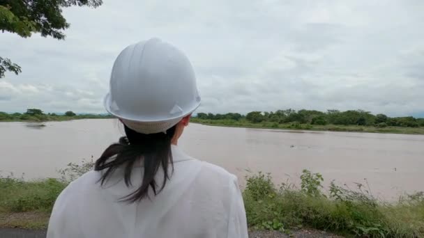 設計図を持っている女性技術者が川の電化ダムの敷地内を歩き 夜の大雨とダムの容量を超える強い電流の後を探索します クリーンエネルギー — ストック動画
