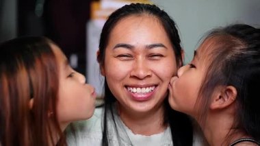 Neşeli bir Asyalı anne gülümserken iki kızı da evde onu yanaklarından öpüyor. İmaj aile sevgisini, sevgiyi, anneliği, mutluluğu ve ebeveynler ile çocuklar arasındaki bağı temsil ediyor.. 