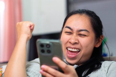 Başarıyı kutlayan Asyalı bir kadının yakın çekimi ve akıllı telefonuna bakarken heyecanlanması. Görüntü mutluluğu, zaferi, iyi haberleri ya da olumlu çevrimiçi tepkiyi temsil ediyor. Kazanan anları, neşeli mesajları ya da duyguları göstermek için mükemmel.