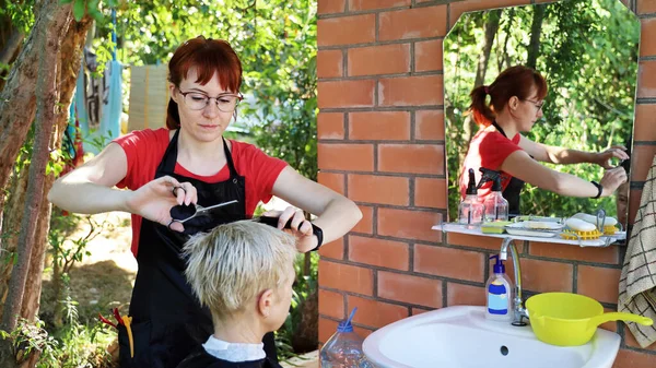 Young hairdresser makes haircut to a woman in front of mirror in the yard outdoors on summer day. During self-isolation, her professional skills come in handy