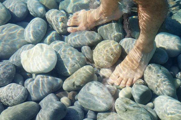 Male bare feet in water on underwater stones in soft focus due to light ripples on water on sunny day