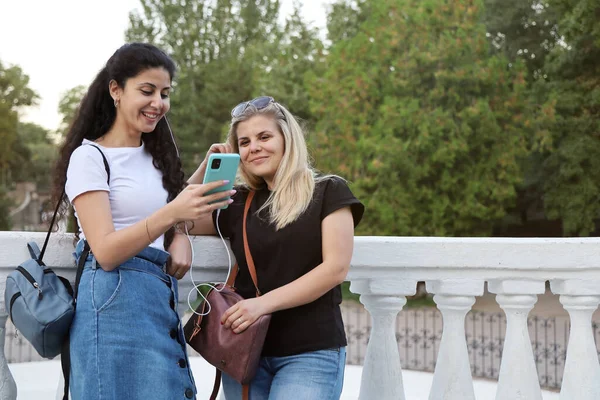 İki gülümseyen bayan arkadaş parkta yan yana durur ve akıllı telefondan aynı kulaklıkla müzik dinlerler.