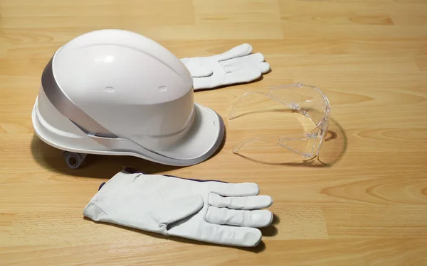 White construction helmet, safety goggles and leather gloves on surface with pattern of wood, close-up. Concept of personal protective equipment and safety for work