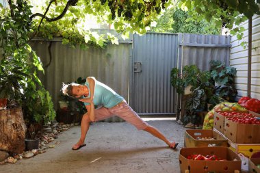 Günlük kıyafet giyen genç kadın, toplanan sebze kutularının istiflendiği avluda otantik bir ortamda egzersiz yapıyor.