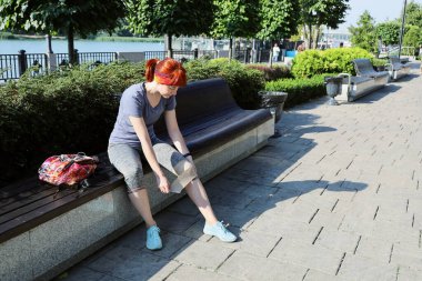 Spor kıyafetli genç kızıl saçlı kadın bankta tek başına oturur ve yaralı dizini elastik bir bandajla bandajlar, güneşli yaz gününde toprak sette.