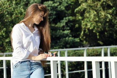 Uzun saçlı, beyaz tişörtlü, kot pantolonlu, gülümseyen genç bir kadın, yeşil arka plan karşısında duruyor, yaz güneşli bir günde fotokopi alanı ile