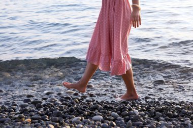 Pembe elbiseli kadın deniz kıyısında çıplak ayakla yürüyor, yakın plan.