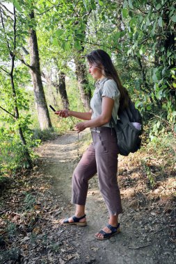 Sırt çantalı genç bir turist orman yolunda duruyor ve yaz günü pusulaya bakıyor.