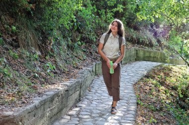 Elinde şişeyle yürüyüş yapan genç bir kadın kayalık yolda yürüyor ve bir yere bakıyor.