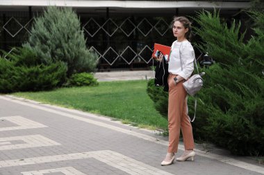 Elinde eğitim malzemeleriyle genç ve güzel bir kadın parkta duruyor ve konsantrasyonuyla uzaklığa bakıyor.