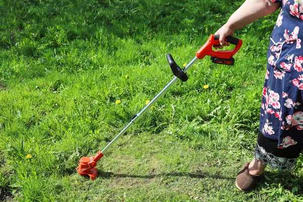 太りすぎの女性の手は草を刈るための電動トリマーを保持しています 緑の草原を背景に 家庭用の便利な軽量ツール — ストック写真