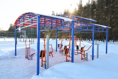 Açık çatı altında spor simülatörleri, kışın dışarıda, güneşli bir günde insanların olmadığı orman sınırına yakın park alanı.